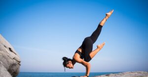 Woman performing a yoga pose on a rocky beach with ocean view and clear blue sky.